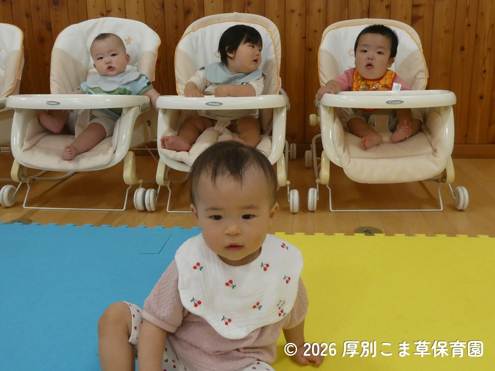 ホールの後方から誕生会の様子を眺めているたんぽぽ組（０歳児）の子どもたちの様子